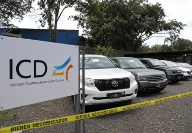 Rótulo del Instituto Costarricense sobre Drogas junto a una fila de automóviles de lujo custodiados por una cinta amarilla de vehículos decomisados al narcotráfico.