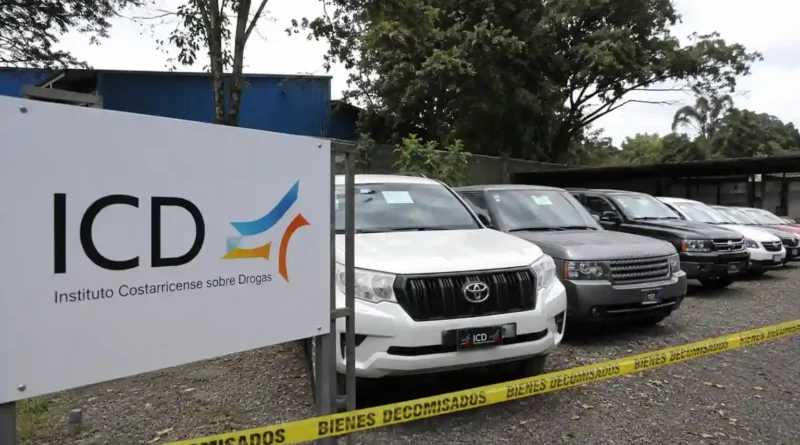 Rótulo del Instituto Costarricense sobre Drogas junto a una fila de automóviles de lujo custodiados por una cinta amarilla de vehículos decomisados al narcotráfico.