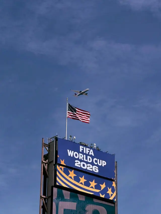 Cartel publicitario oficial de la FIFA World Cup 2026 con la bandera de Estados Unidos ondeando arriba y un avión sobrevolando el cielo azul.